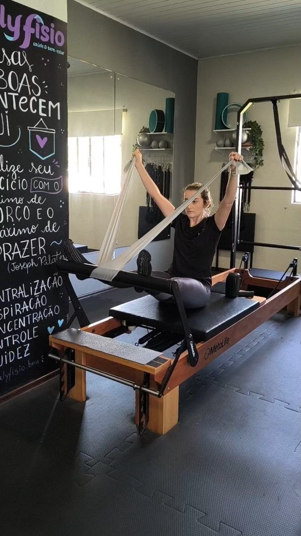 Aluna no reformer da QualyFisio, segurando as alças com os braços estendidos, ao lado da lousa da sala